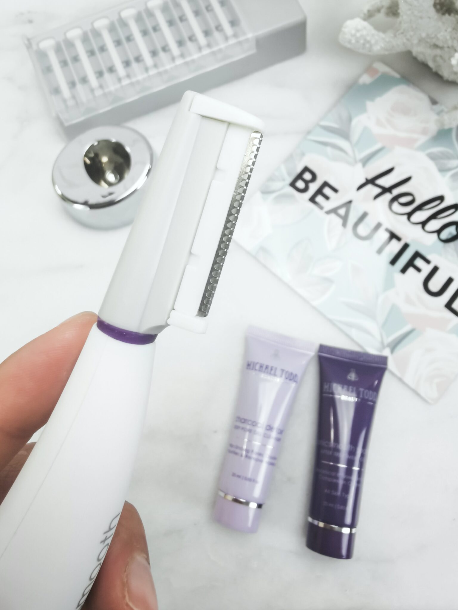 Close up of Sonicsmooth blade, cleanser, treatment gel, charging stand, replacement, blades, a sign that says, "Hello Beautiful," and a white coral is pictured. 