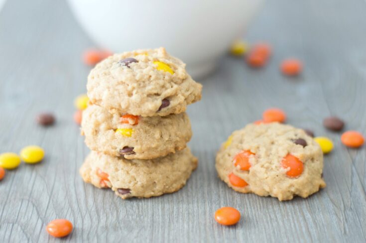 A bowl full of Reese's Pieces Peanut Butter Oatmeal cookies with 3 cookies on the table surrounded by Reese's Pieces. They sit on a grey table.