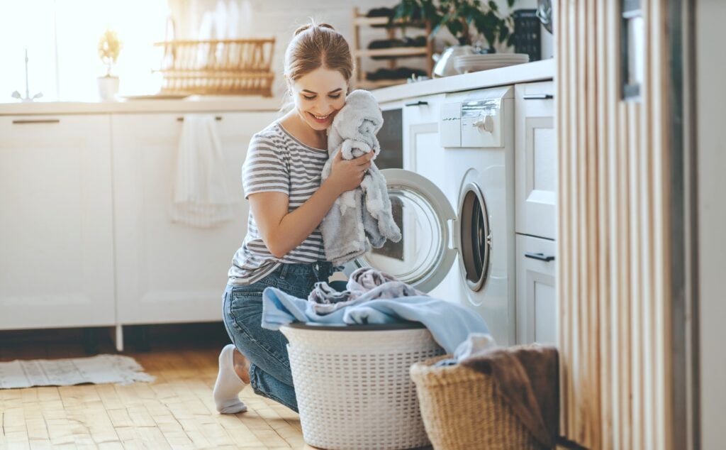 Woman takes out laundry from front facing washing machine. She smiles and holds a clean towel to her face while she loads the basket. 