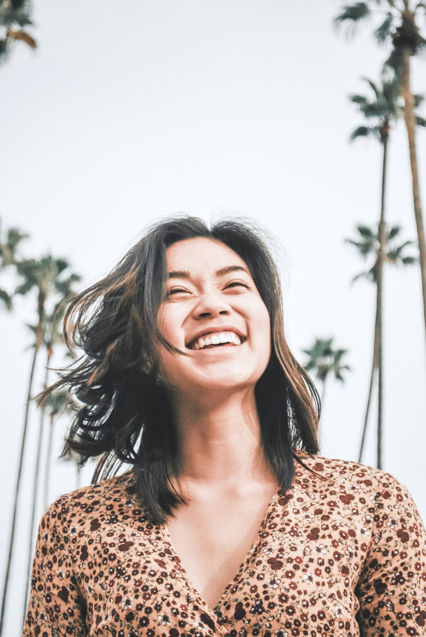 A woman smiles and looks up to the sky. There are palm trees behind her. A great smile is one of the beauty and fashion trends - summer 2020.