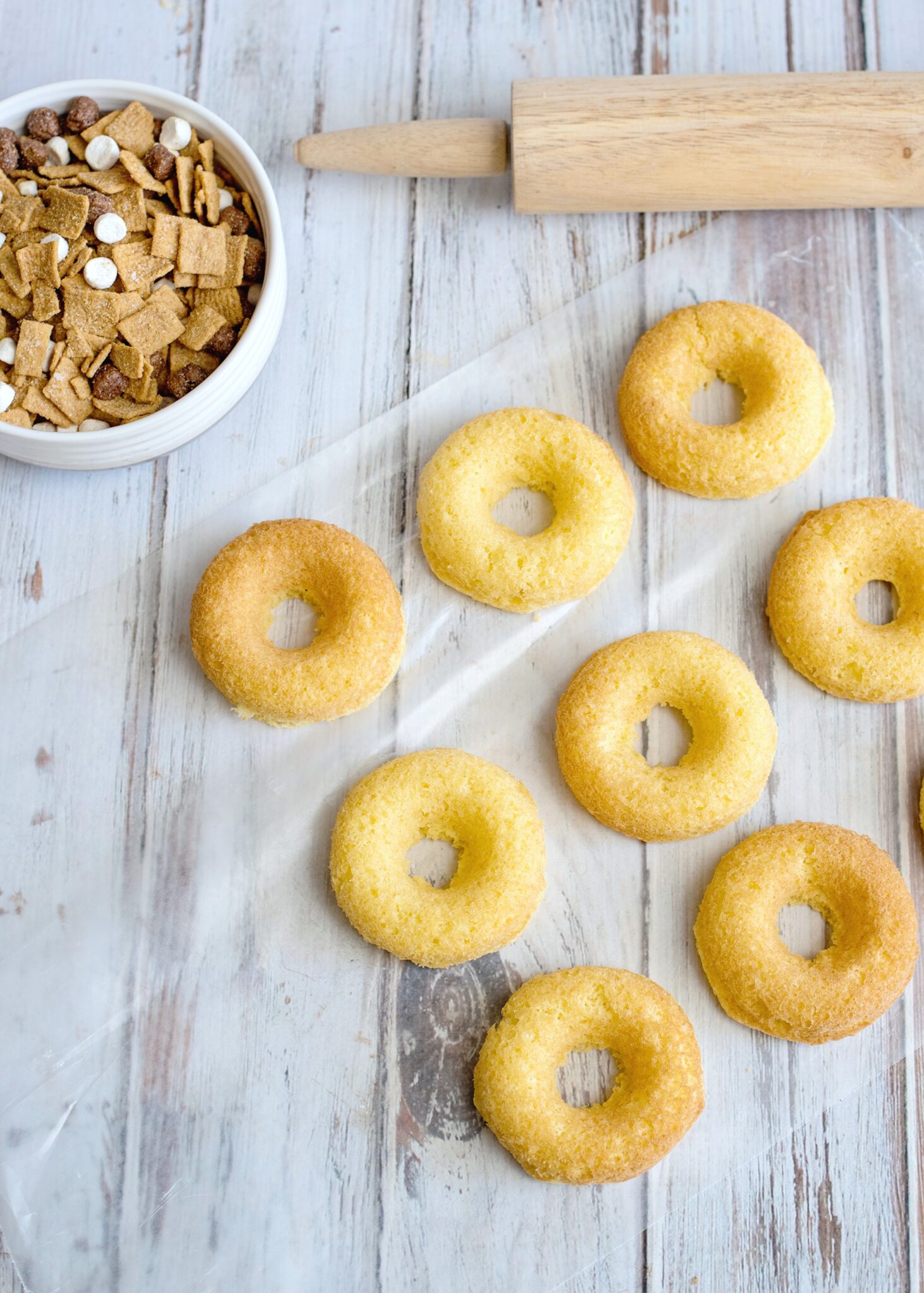 Baked donuts cooling on parchment paper. A rolling pin and bowl of cereal is shown.