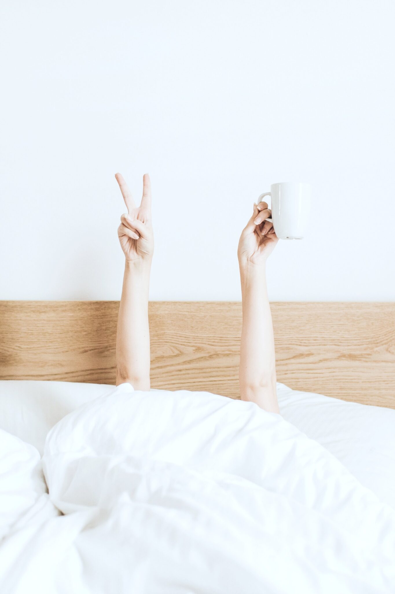 A woman hides under her sheets in bed and raises a peace sign and a coffee mug.