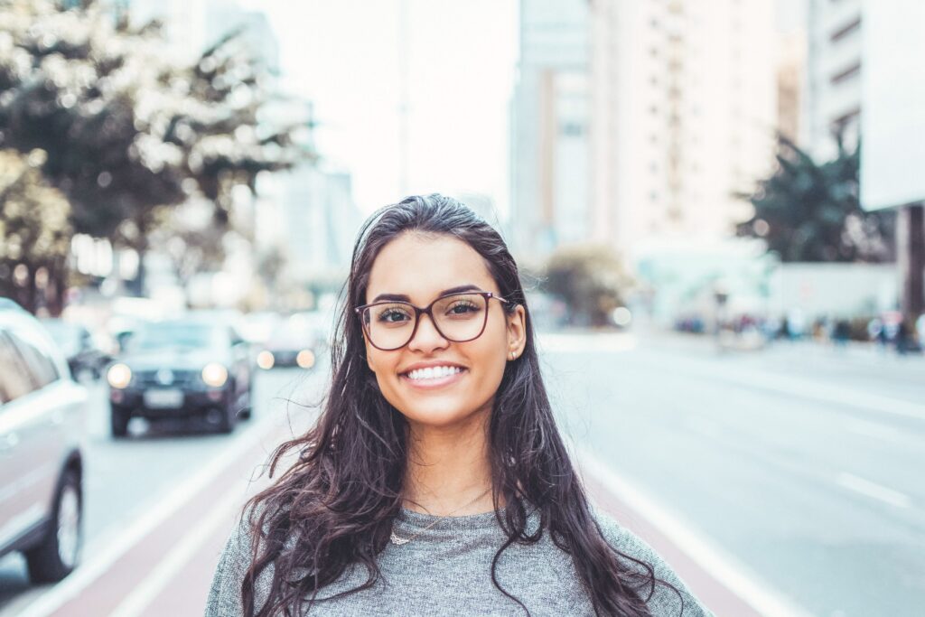A woman stands on a busy street and smiles. She is wearing glasses and has a beautiful smile. 