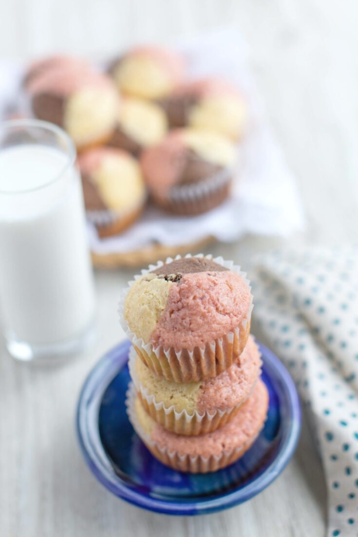 A stack of Neapolitan Ice Cream Muffins stacked on a plate. Behind it is a glass of milk and a full plate of muffins.