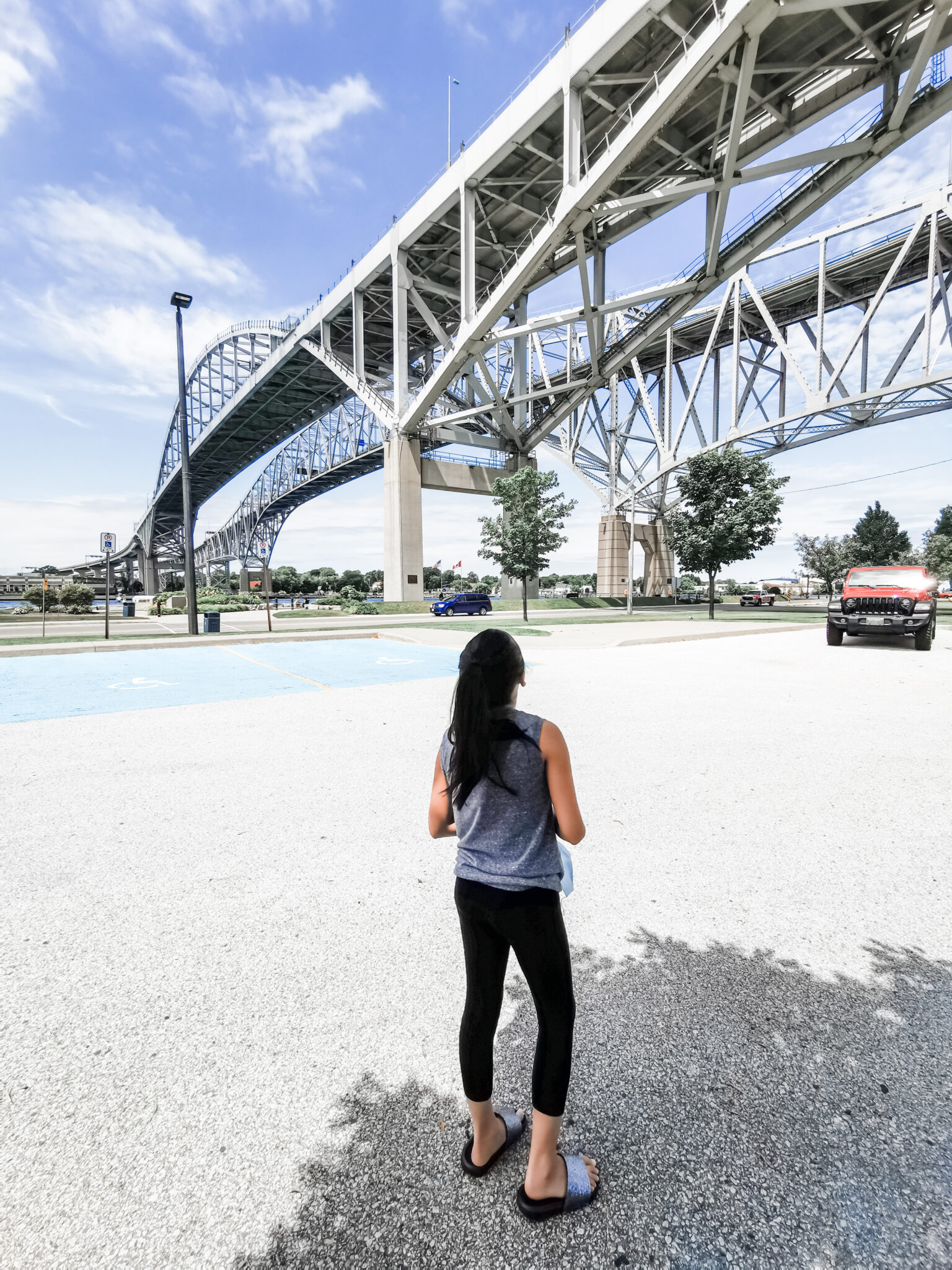 Mimi looks up at the Bluewater Bridge, while a red Jeep is in the background. This is part of roadtrippin' with ProClip USA Jeep Accessories.