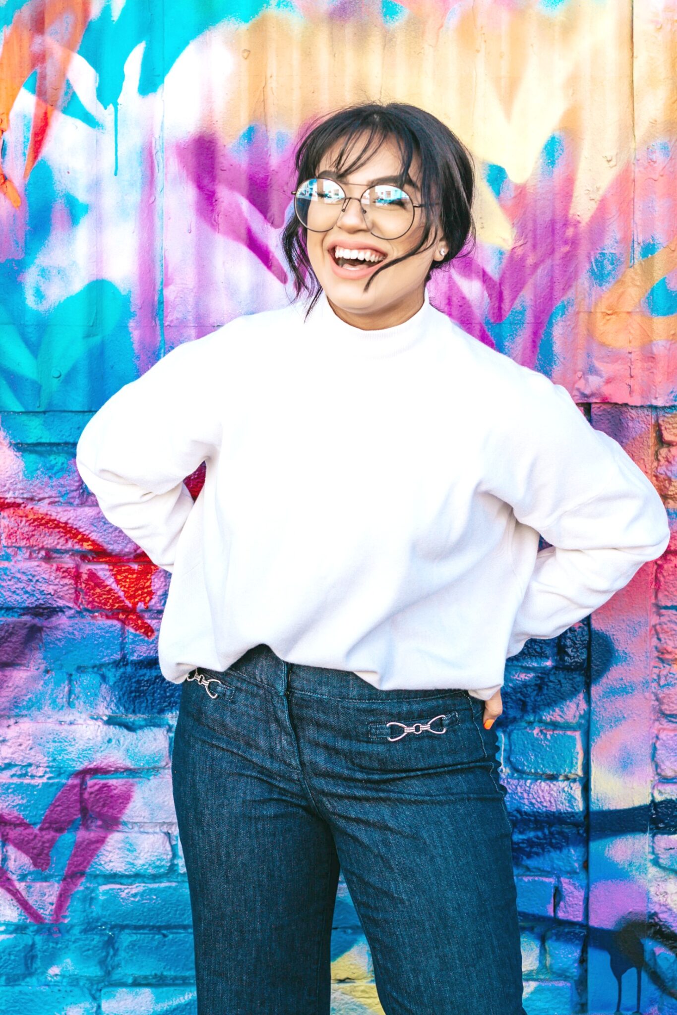 Beautiful Hispanic woman smiling and laughing, she is wearing glasses and stands in front of a colourful wall. 