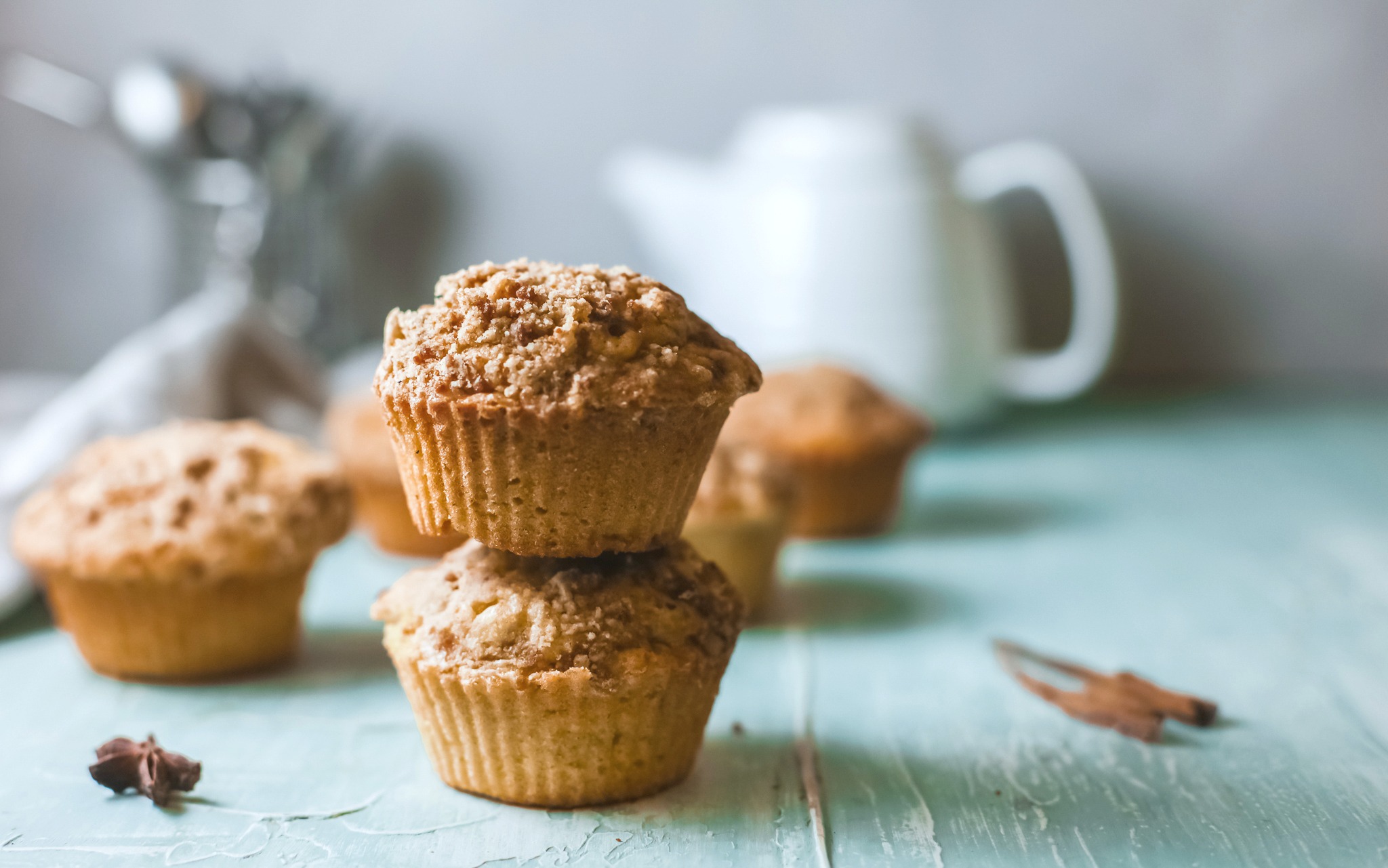 A stack of two pumpkin streusel muffins, there are more muffins behind it with a tea pot and utensils.
