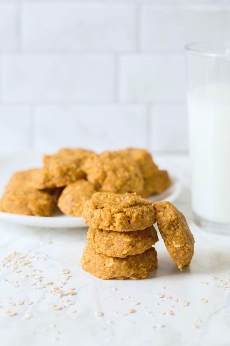 A plate of no-bake pumpkin oatmeal cookies and a stack of cookies on the side. There is a glass of milk against a white background.
