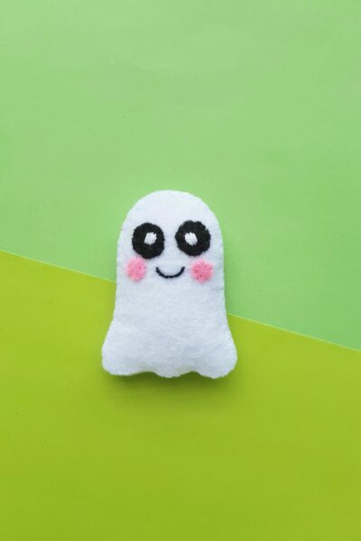 A cute little felt ghost plush sits on a light and dark green background. It has rosy cheeks and cute bright eyes.