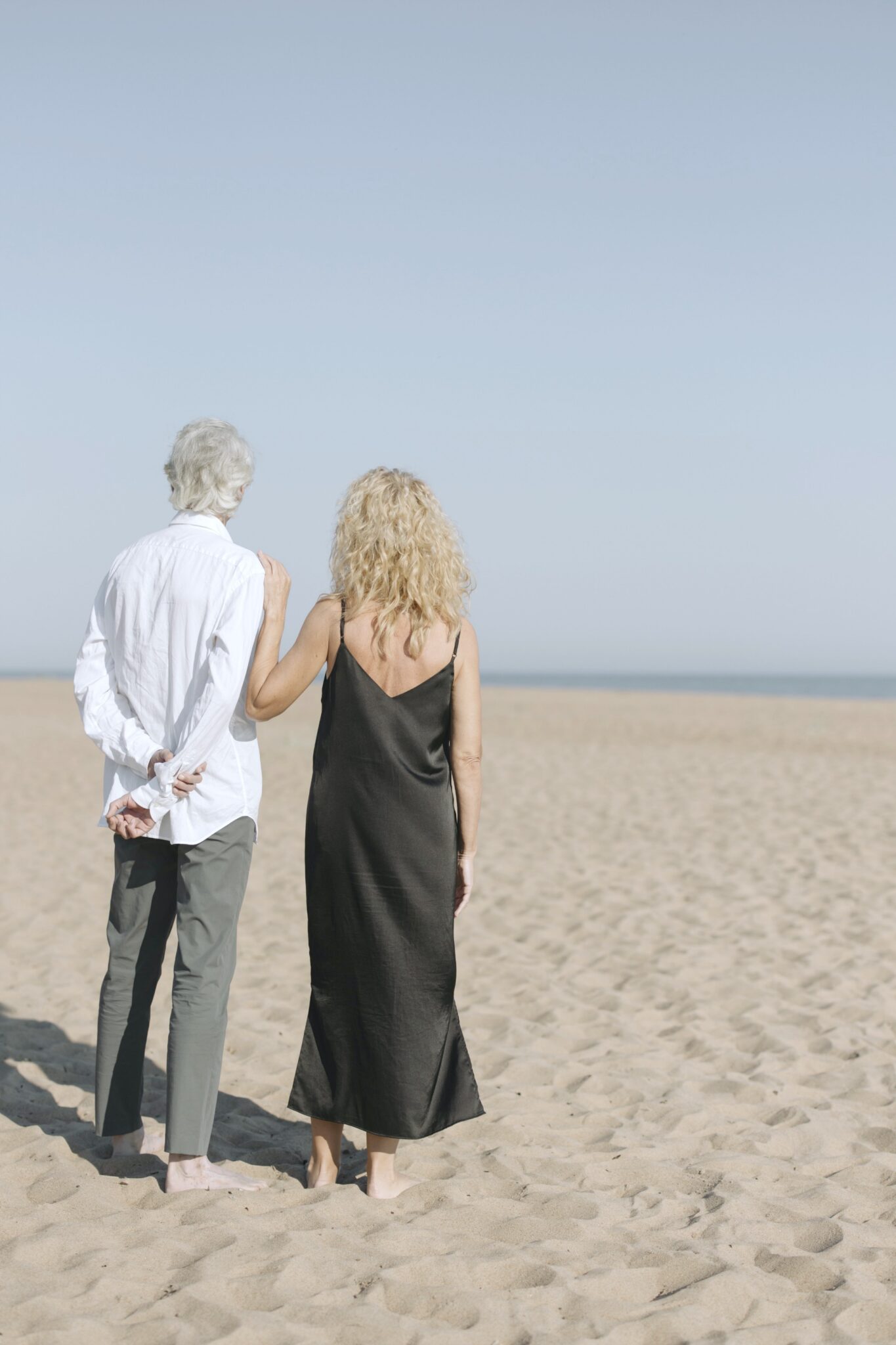 An older couple stand on a beach and look beyond the horizon. The woman has an arm on his shoulder.