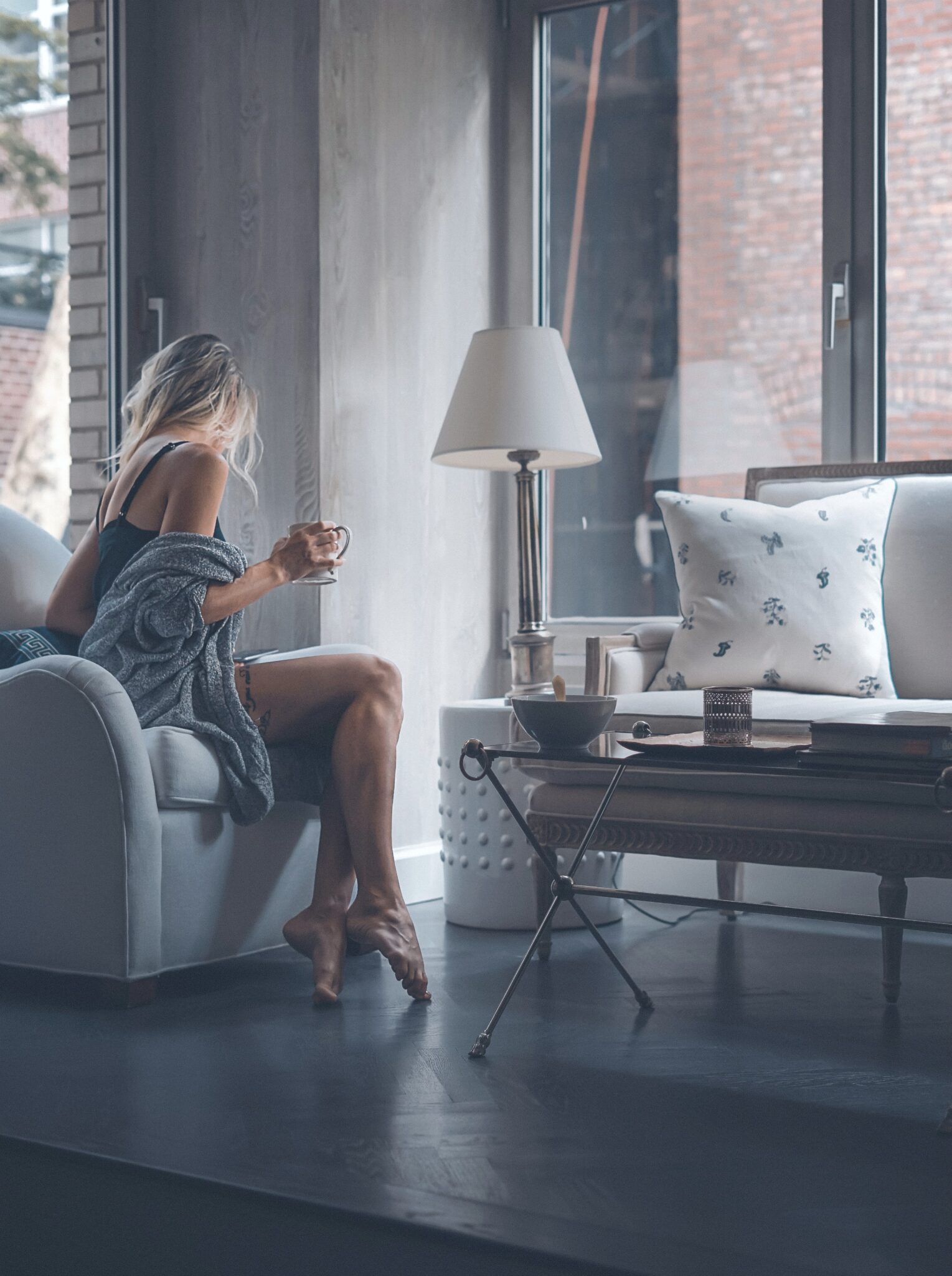 A blonde woman sits on a couch in her New York apartment while holding a mug.