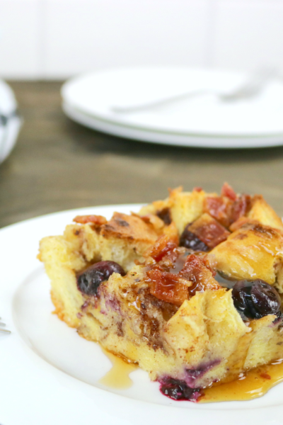A pan of Blueberry Bacon Bread pudding is on a small plate, maple syrup is drizzled on top with a fork.