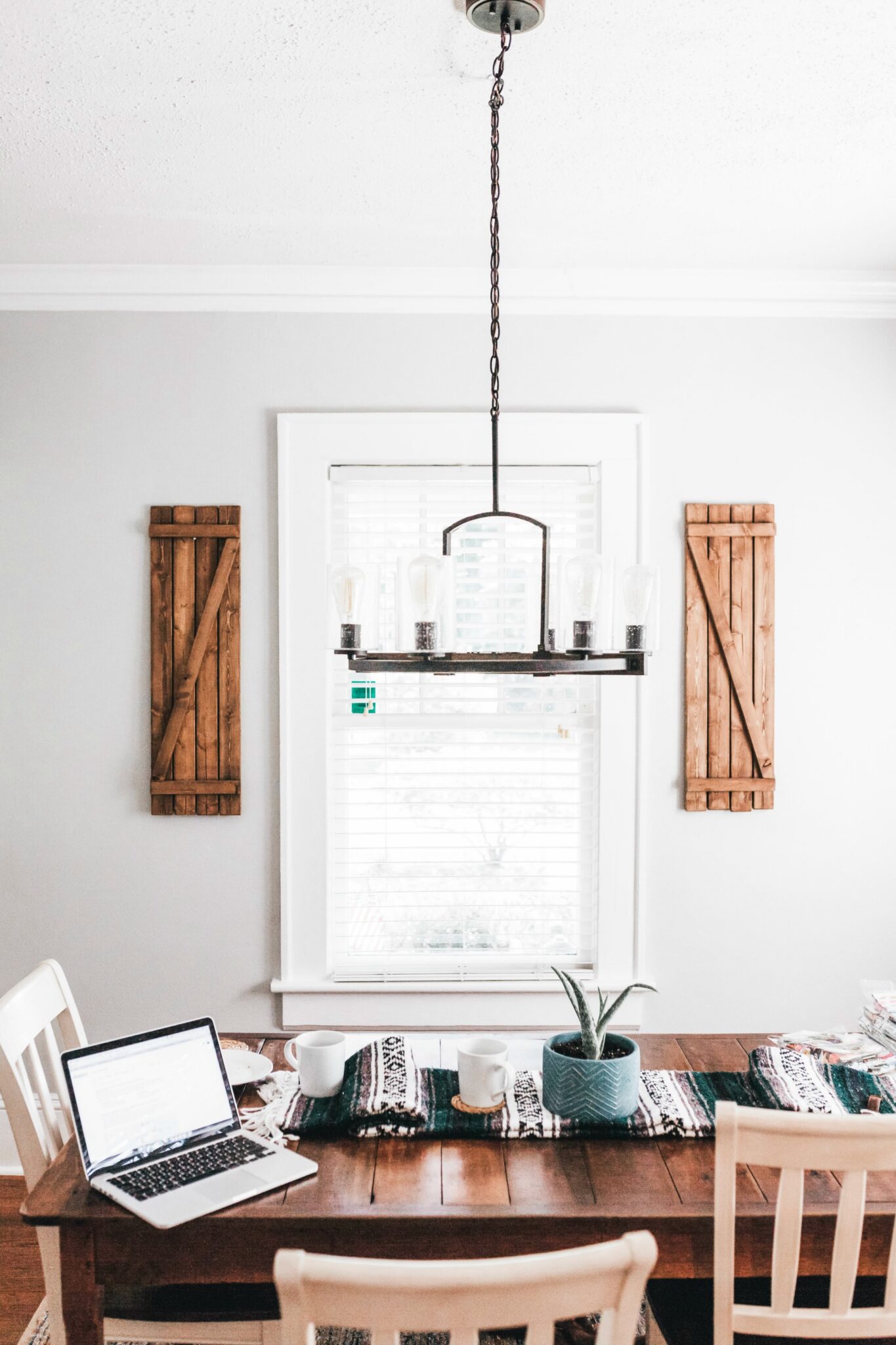 A beautiful farmhouse kitchen showing a bright window with blinds. A chandelier hangs and below is a makeshift office. 