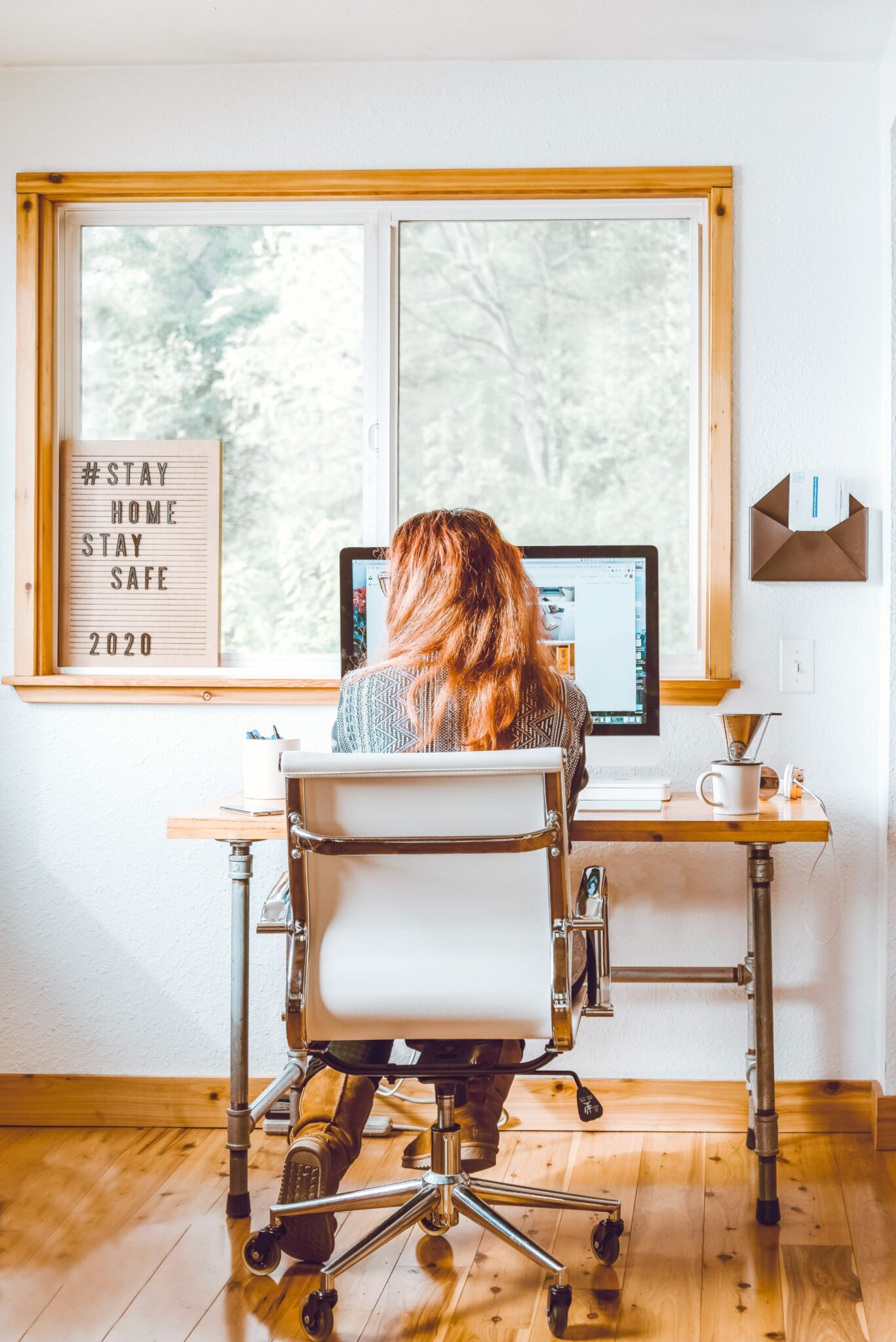 Woman sits at a desk in front of a window. She is working on a computer and her back is facing the camera.