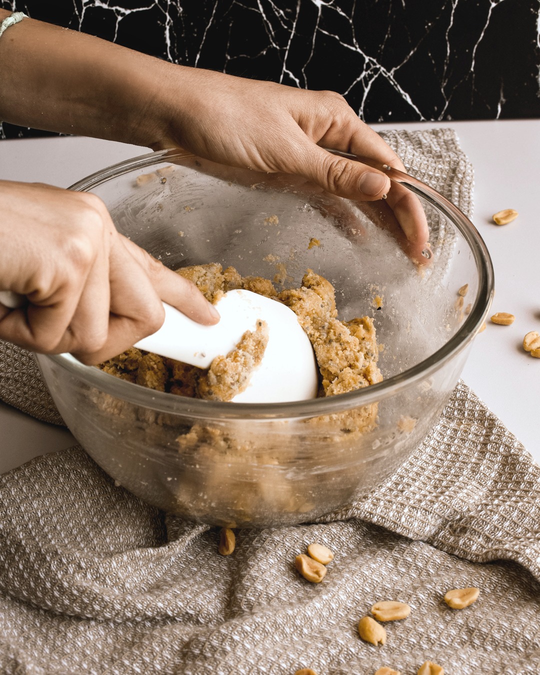 A hand stirs and mixes ingredients together in a medium mixing bowl. It sits on top of a brown napkin and peanuts surround it.