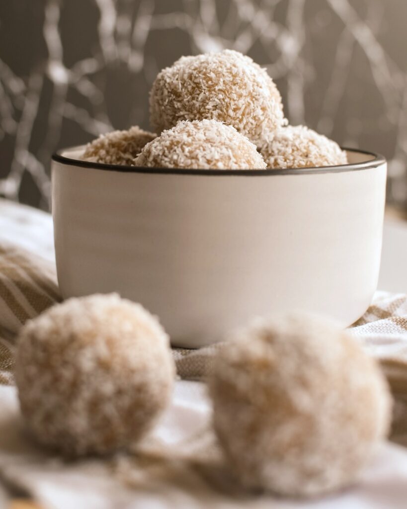 A bowl is filled with peanut butter coconut balls. They are in focus while two protein balls are out of focus in front of the bowl. 