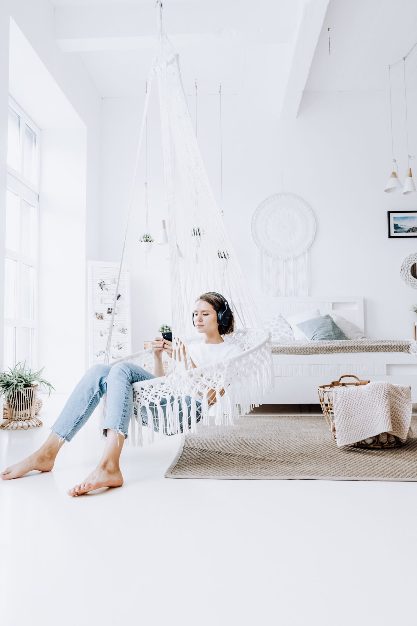 A woman in a bright white room with macramé sits in a macramé swing, listening to something on her phone with headphones. A great place to enjoy Christian audio books.