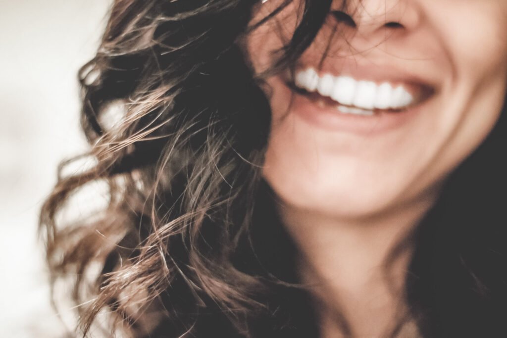 A close up of a woman's face and mouth, she smiles a bright smile. She has dark curly hair. The rest of her face is not seen. This article covers all you need to know about getting a bright smile. 