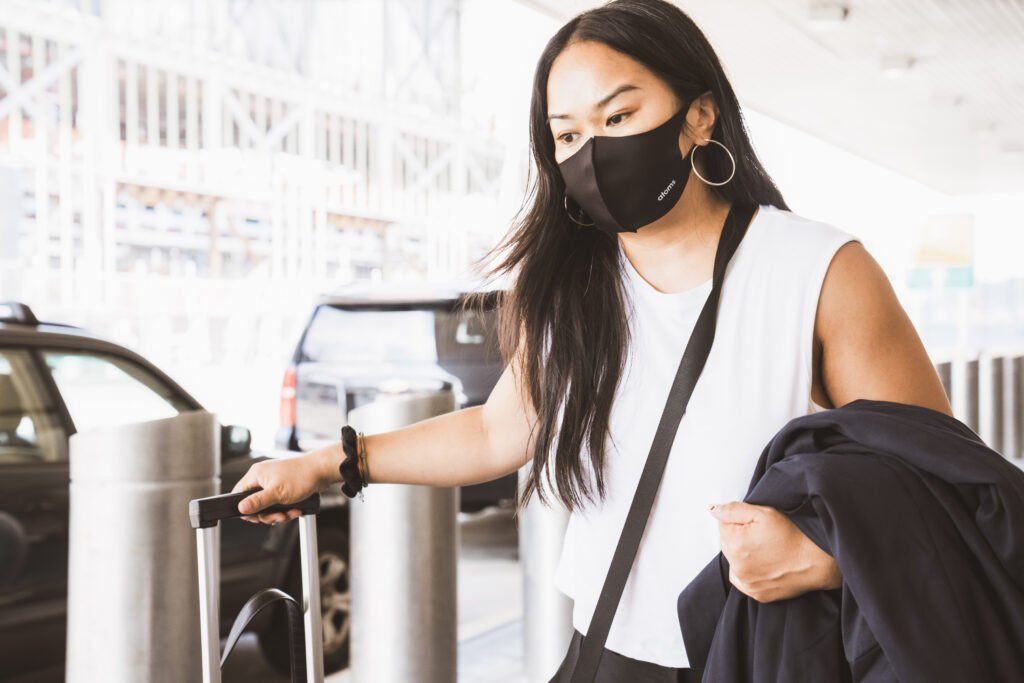 A woman wears a black mask, holds a jacket, and drags a travel luggage. This article covers tips for Travelling in the United States in 2021.

