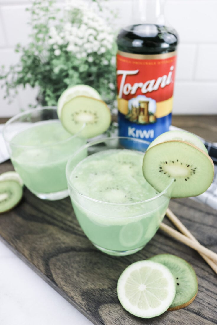 A top view of two cocktails on a board, they're surrounded my kiwi and lime slices and Torani Kiwi Syrup.