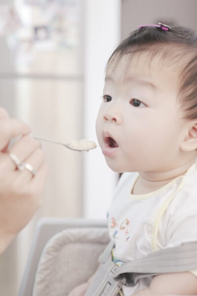 Baby opens his mouth and is being fed with a spoon. This article covers solid foods to avoid feeing your baby within the first year.