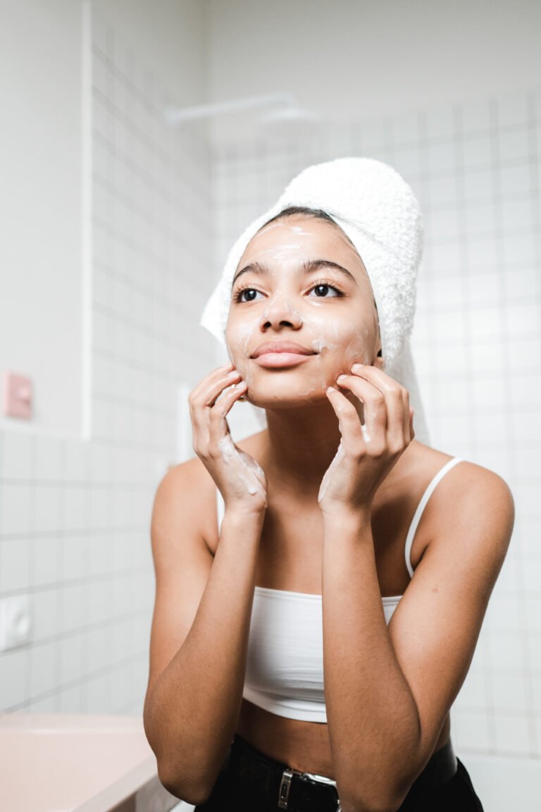 Girl puts on skin care products and looks in mirror. She has a towel on her head. This article covers how to wear make up when you have acne.