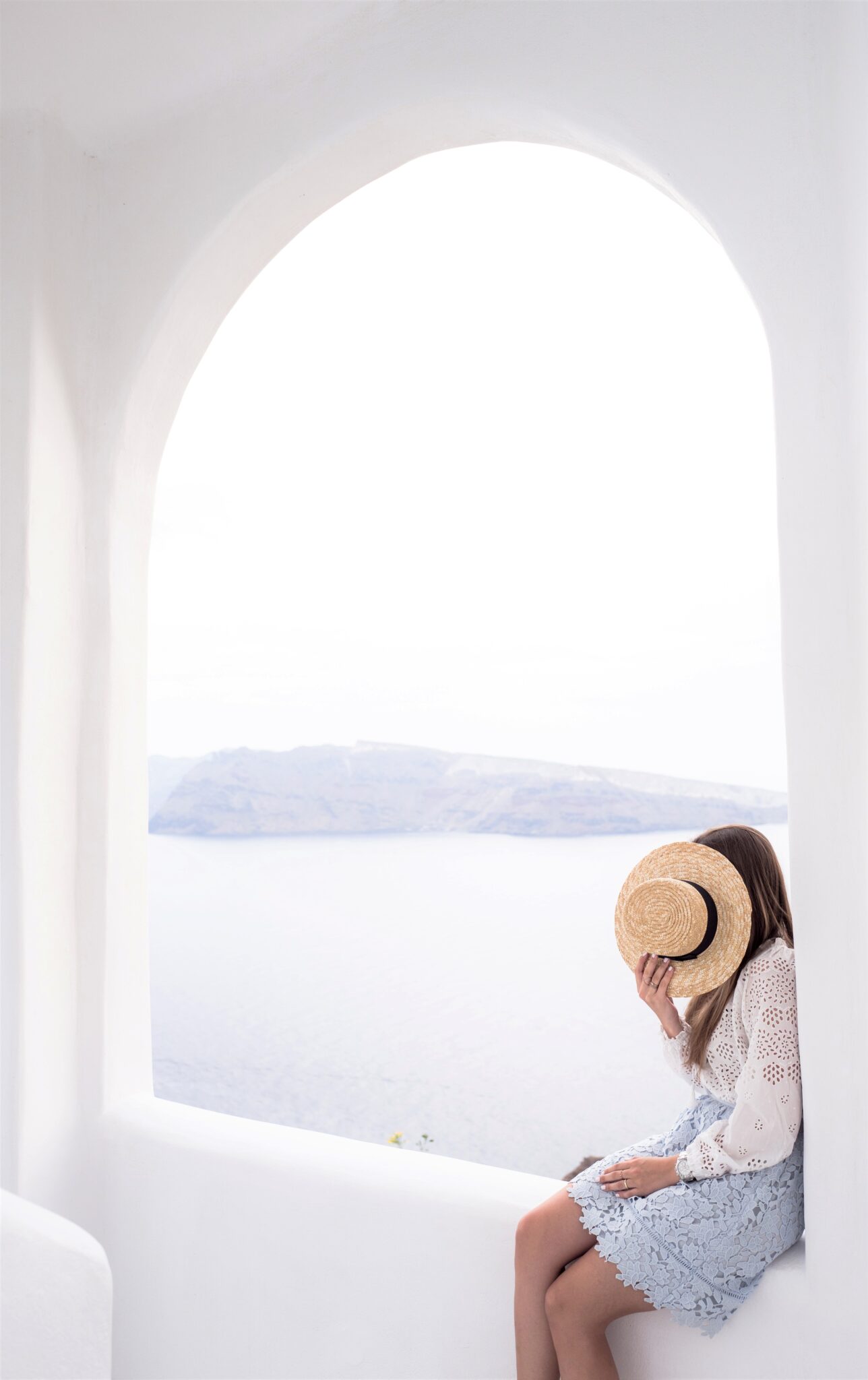A woman sits in a white walkway in Greece. She covers her face with a hat. This article covers 7 Safety Tips to Travel Safe Everywhere.