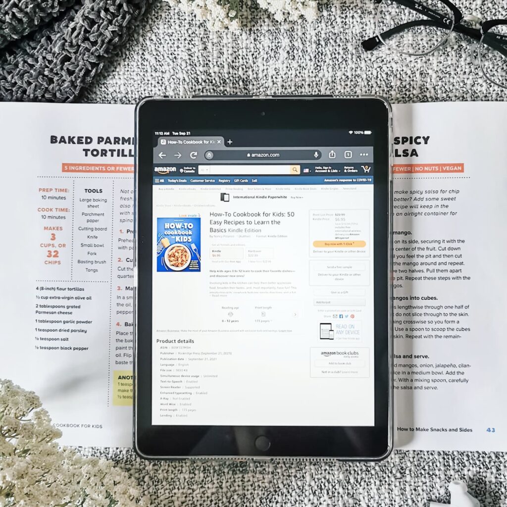 An iPad is placed on top of a cookbook, the cookbook is the "How-To Cookbook for Kids." Surrounding it is a pair of glasses and ear buds.