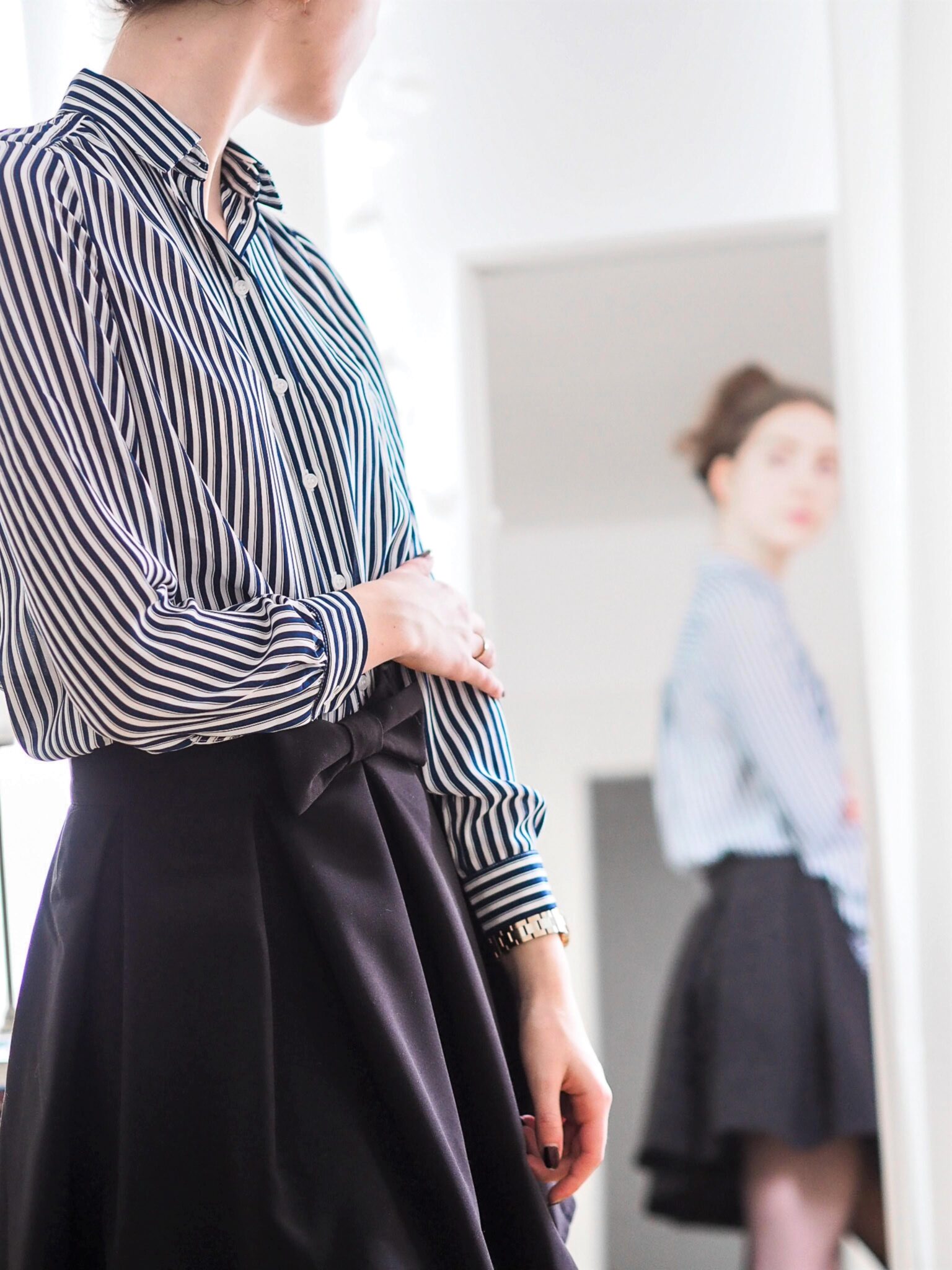 Woman looks at herself in the mirror. She is wearing a dress shirt and skirt. This article covers the best skirt styles to compliment your body shape. 