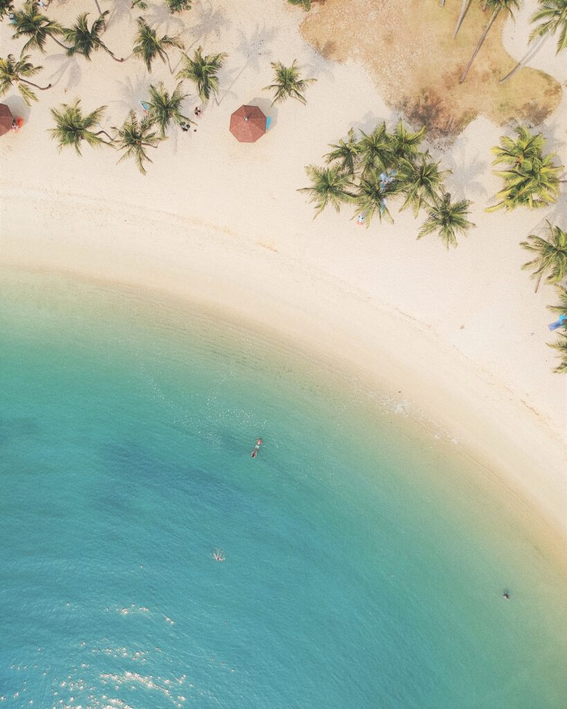 A bird's eye view of a beach, there is a person floating in the water. This article covers reasons to book a long-term holiday.
