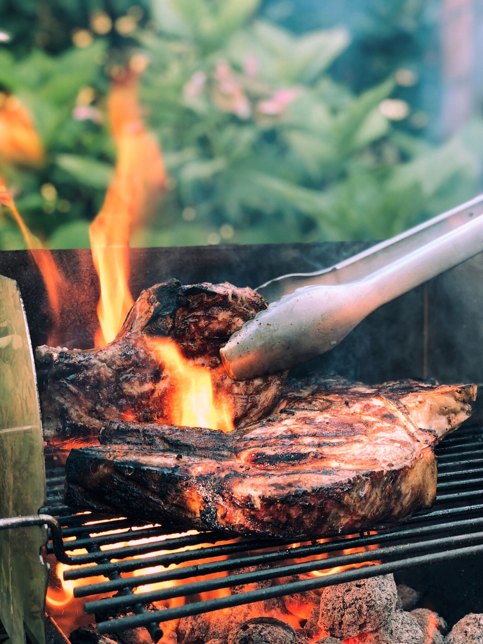 Steak being BBQ'd on a charcoal BBQ. The fire is rising high and a pair of tongs are being used to flip the meat. This article covers how to host the perfect family BBQ.