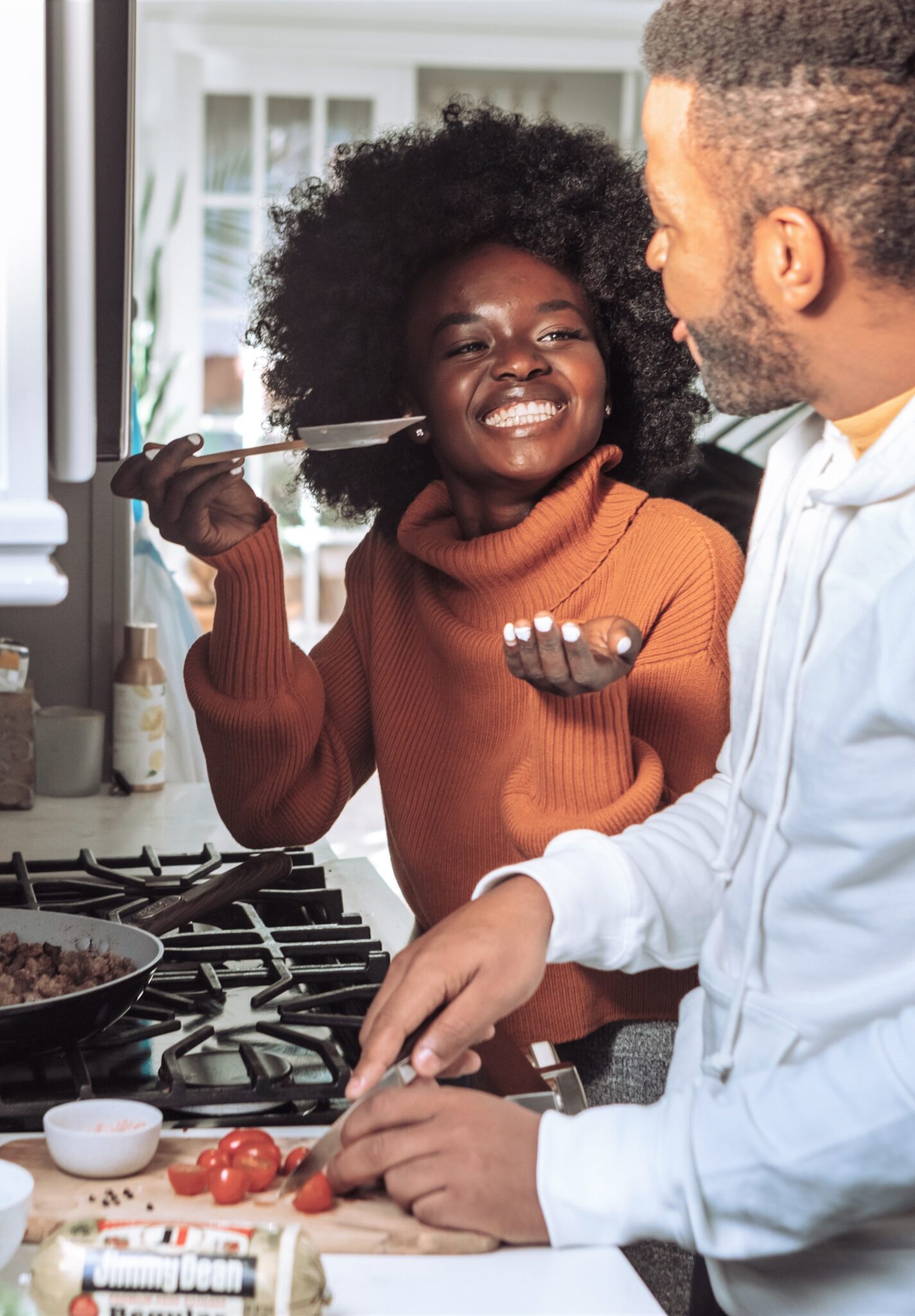 A young couple cook together and the woman is giving her partner a taste of their food.