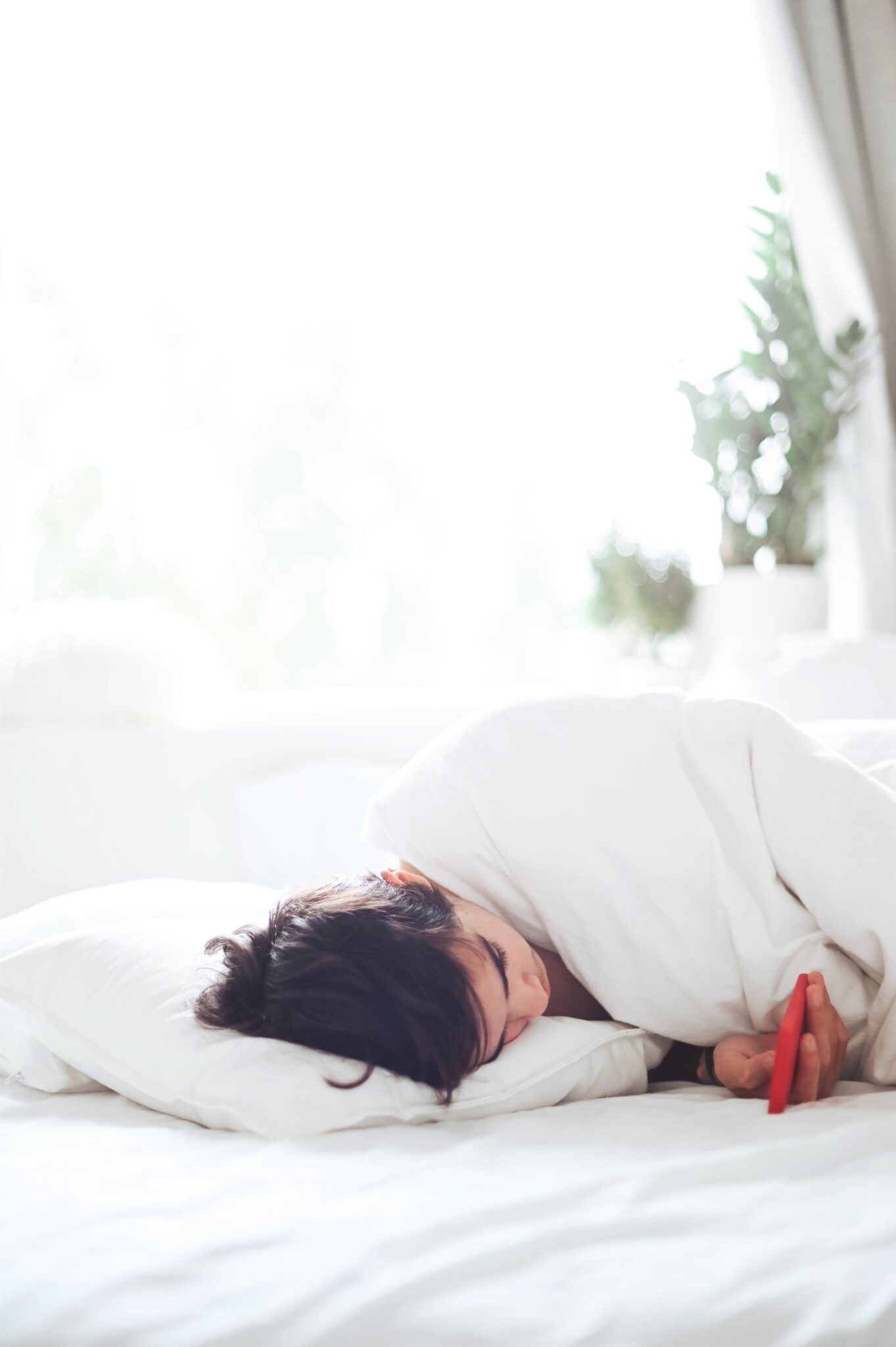 Woman on her phone laying down in bed. The sheets are white and there is a plant behind her. This article covers why you need a wedge pillow.