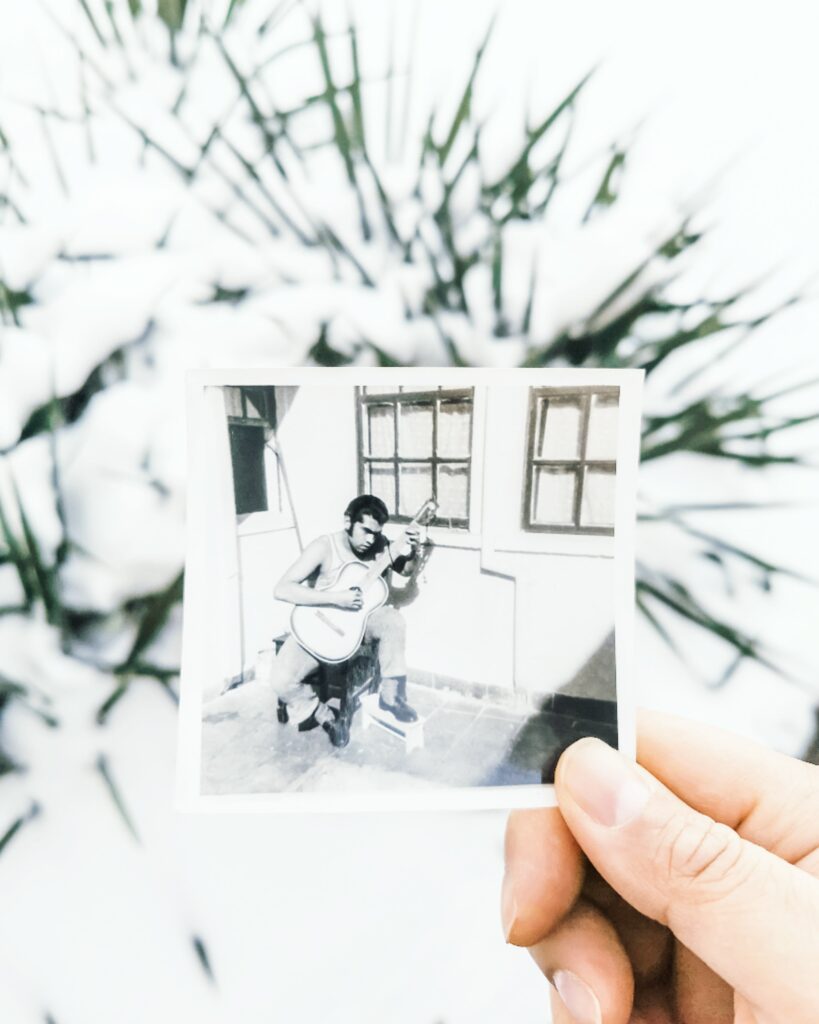 Nancy's hand holds up a vintage polaroid of her dad outside playing a guitar. It's held outside in front of an evergreen bush.