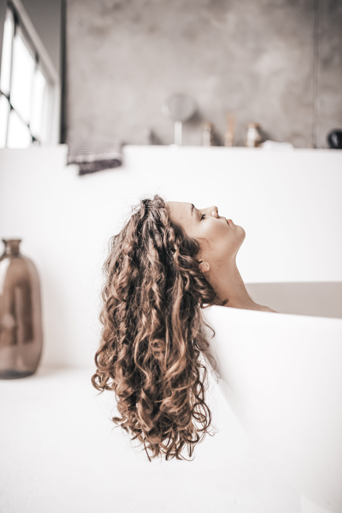 A woman is lying in her tub, eyes closed, and relaxed. Her beautiful, healthy, curly hair is hanging over the tub. This article covers the ultimate guide to your hair care routine. 