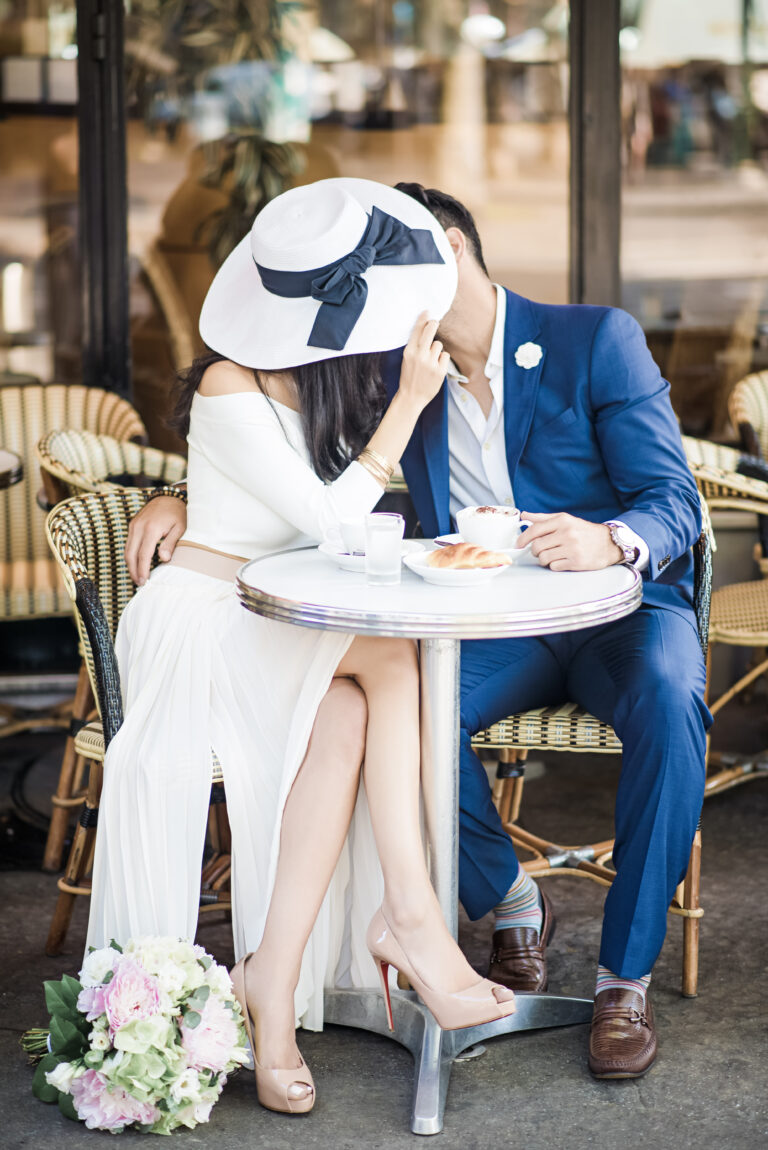 A man and woman kiss, their faces are hidden by the woman's hat. They sit in a bistro. This article covers how to plan a romantic meal for two.