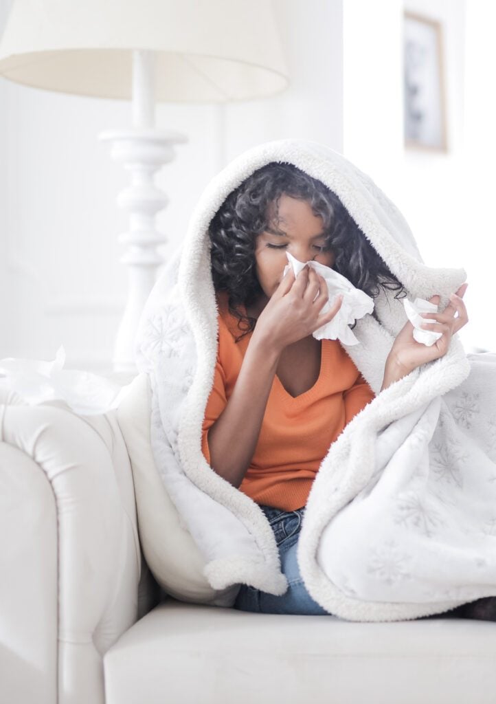 Girl sneezing and blowing her nose with tissues. She is wrapped in a blanket. This article covers the best ways to prepare your house for allergy season.