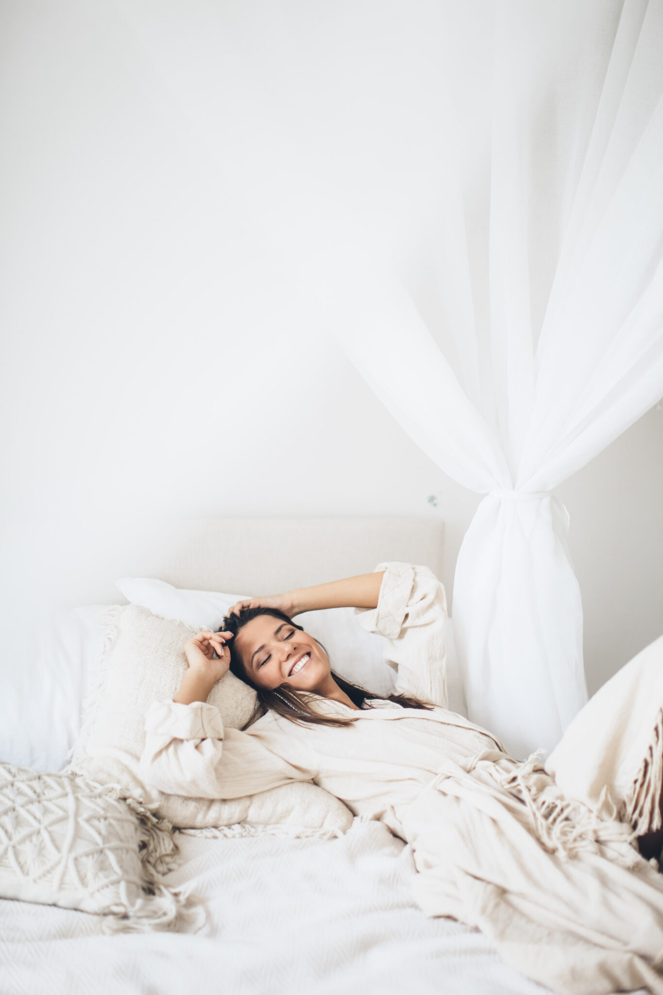 A woman lies in her bed on white sheets, she is wearing neutral pajamas and is closing her eyes and smiling. This article covers tips to achieve happiness. 