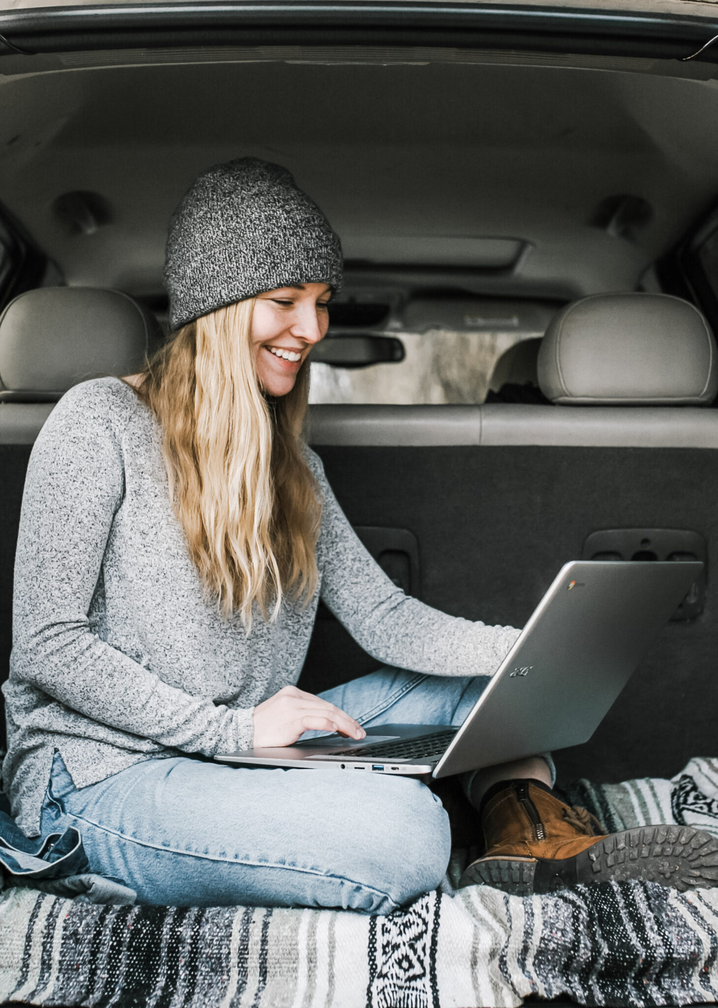 A girl wearing a beanie smiles and uses a laptop in the trunk of an SUV. This article covers ways to combine online studying and traveling in 2022.