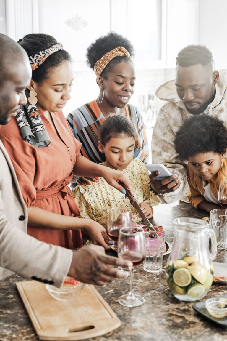 A family gathers around the kitchen cutting a cake. They are huddled around smiling and taking pictures. This article covers how to make the kitchen the heart of your home.