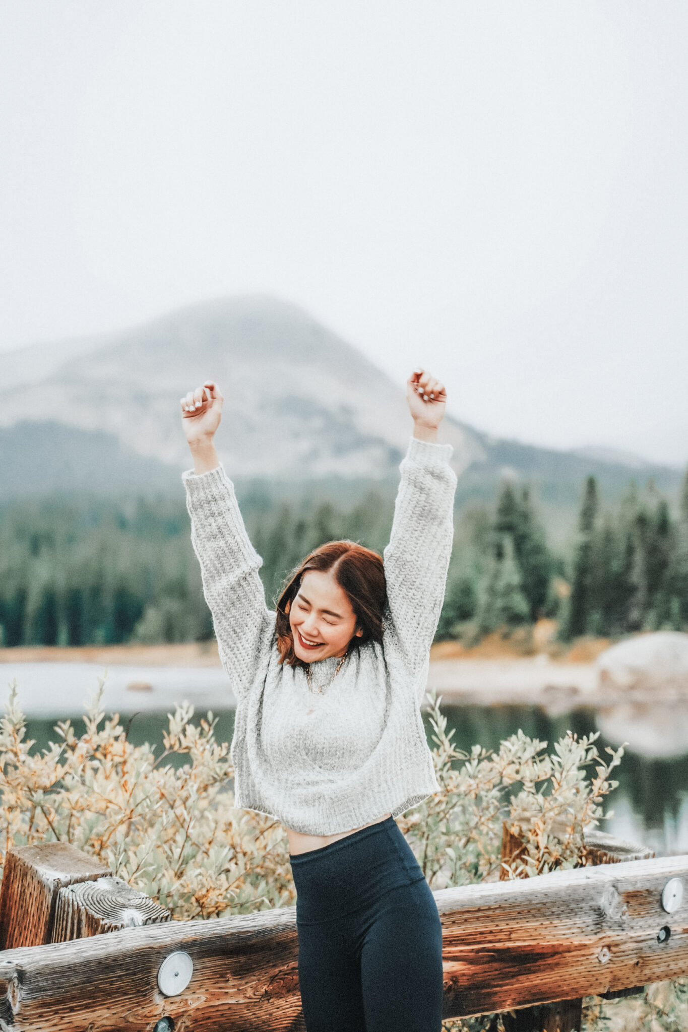 A woman throws her hands in the air, closes her eyes, and smiles. She is outside by a lake surrounded by nature. This article covers style tips for staying warm without looking bulky.