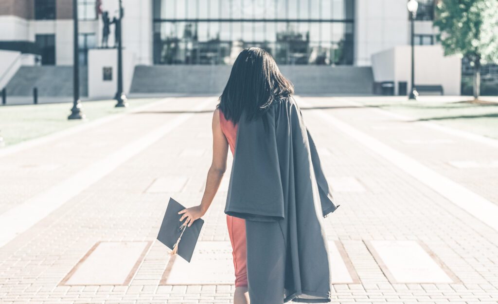 A woman walks away and towards a school building, she has a gown on one shoulder and a grad cap in one hand. Her back faces us. This article covers College Degree Myths.