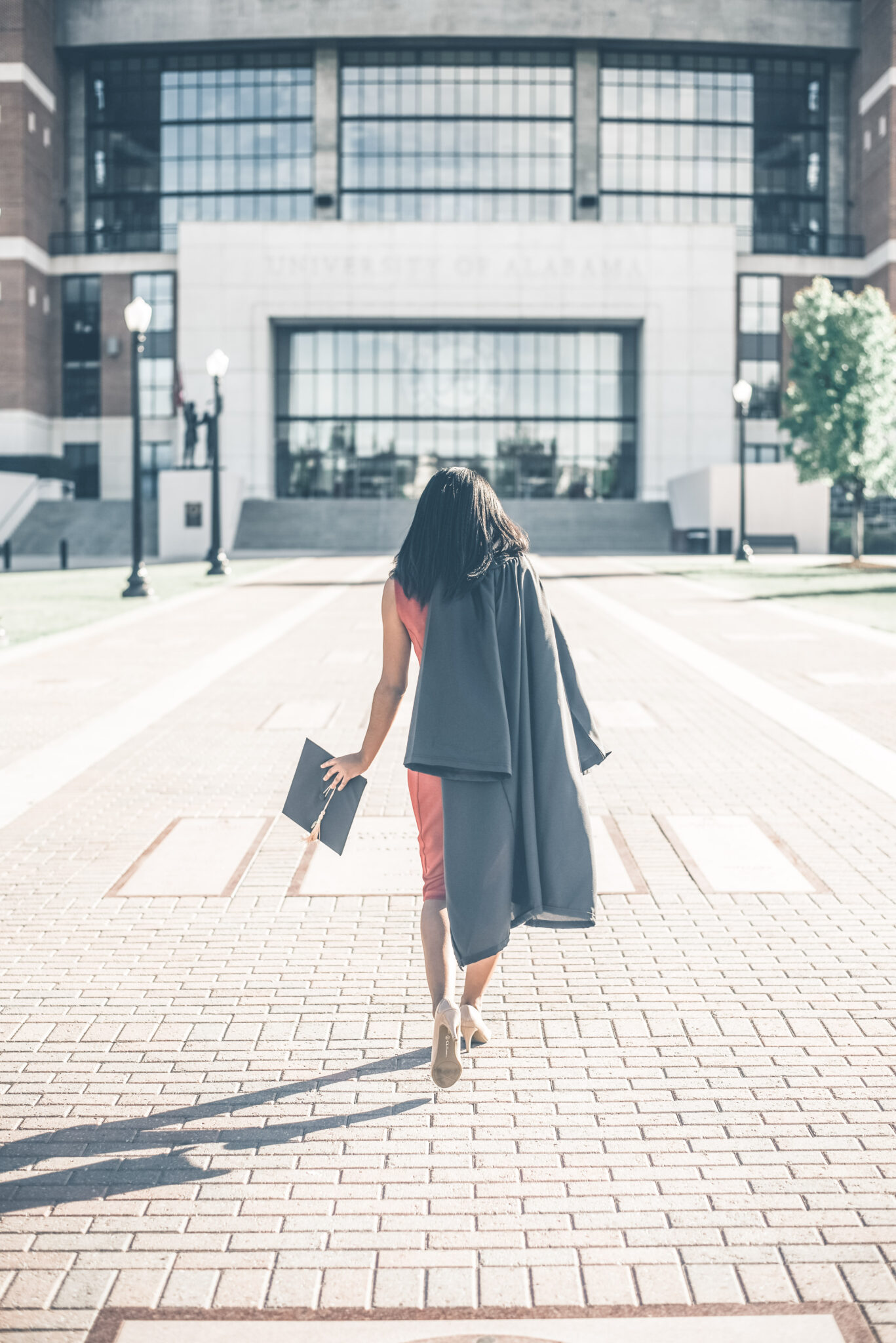 A woman walks away and towards a school building, she has a gown on one shoulder and a grad cap in one hand. Her back faces us. This article covers College Degree Myths. 