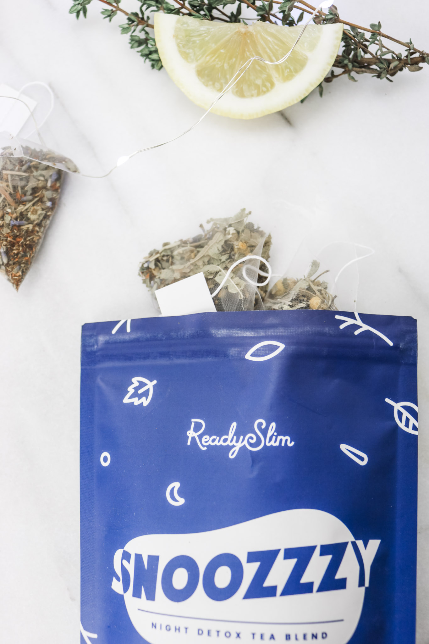 A ReadySlim bag is open with tea bags falling out. There is a slice of lemon and thyme and fairy lights surrounding it.