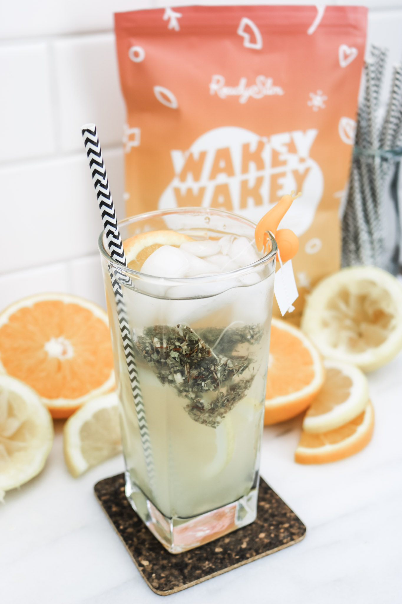 A tall glass of Wakey Wakey tea is shown with ingredients used to make it surrounding it. A bag of Wakey Wakey is behind it. 