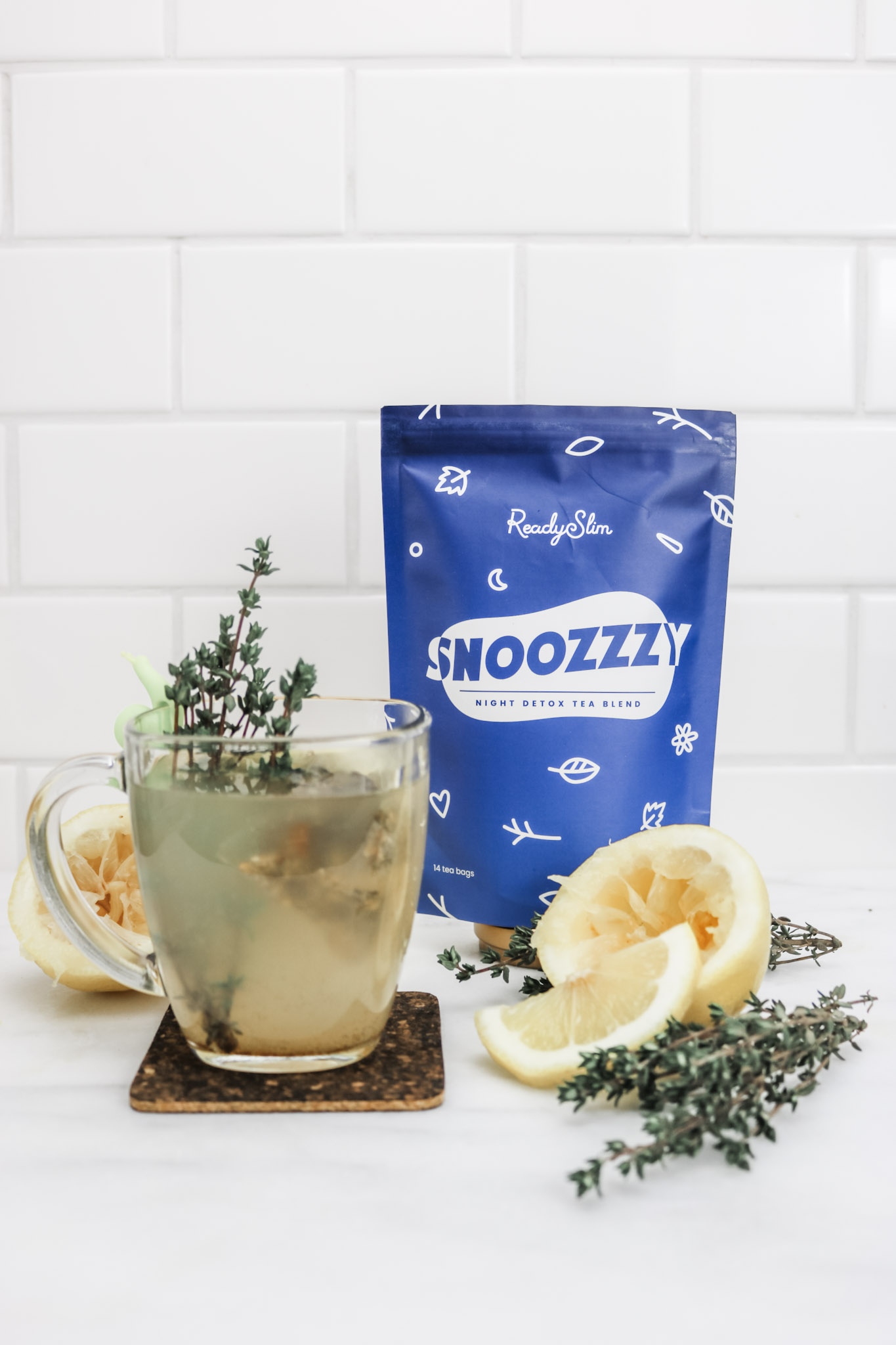 A mug of Snoozzzy tea is shown with ingredients used to make it surrounding it. A bag of Snoozzzy is behind it. 