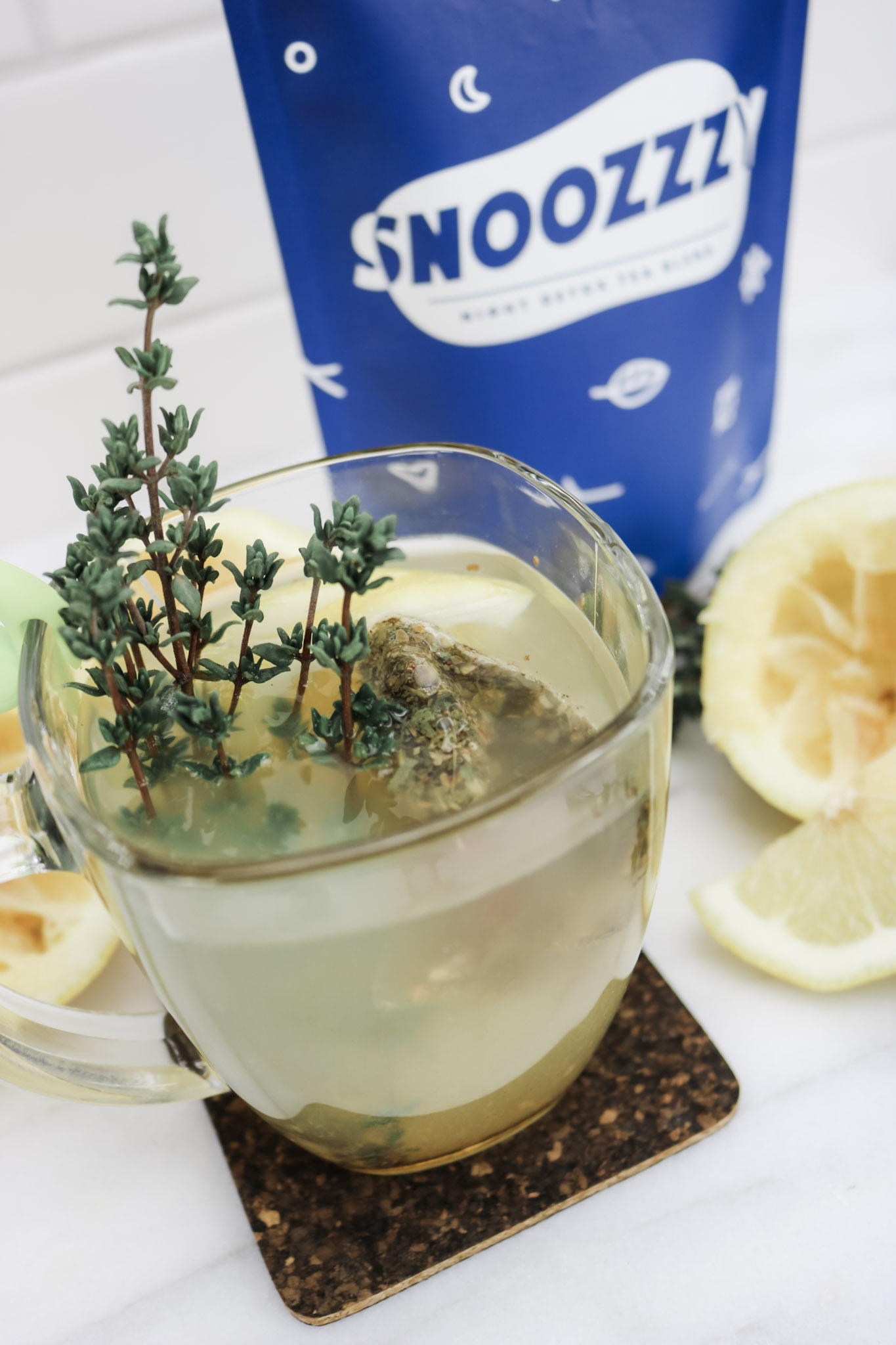A mug of Snoozzzy tea is shown with ingredients used to make it surrounding it. A bag of Snoozzzy is behind it. 