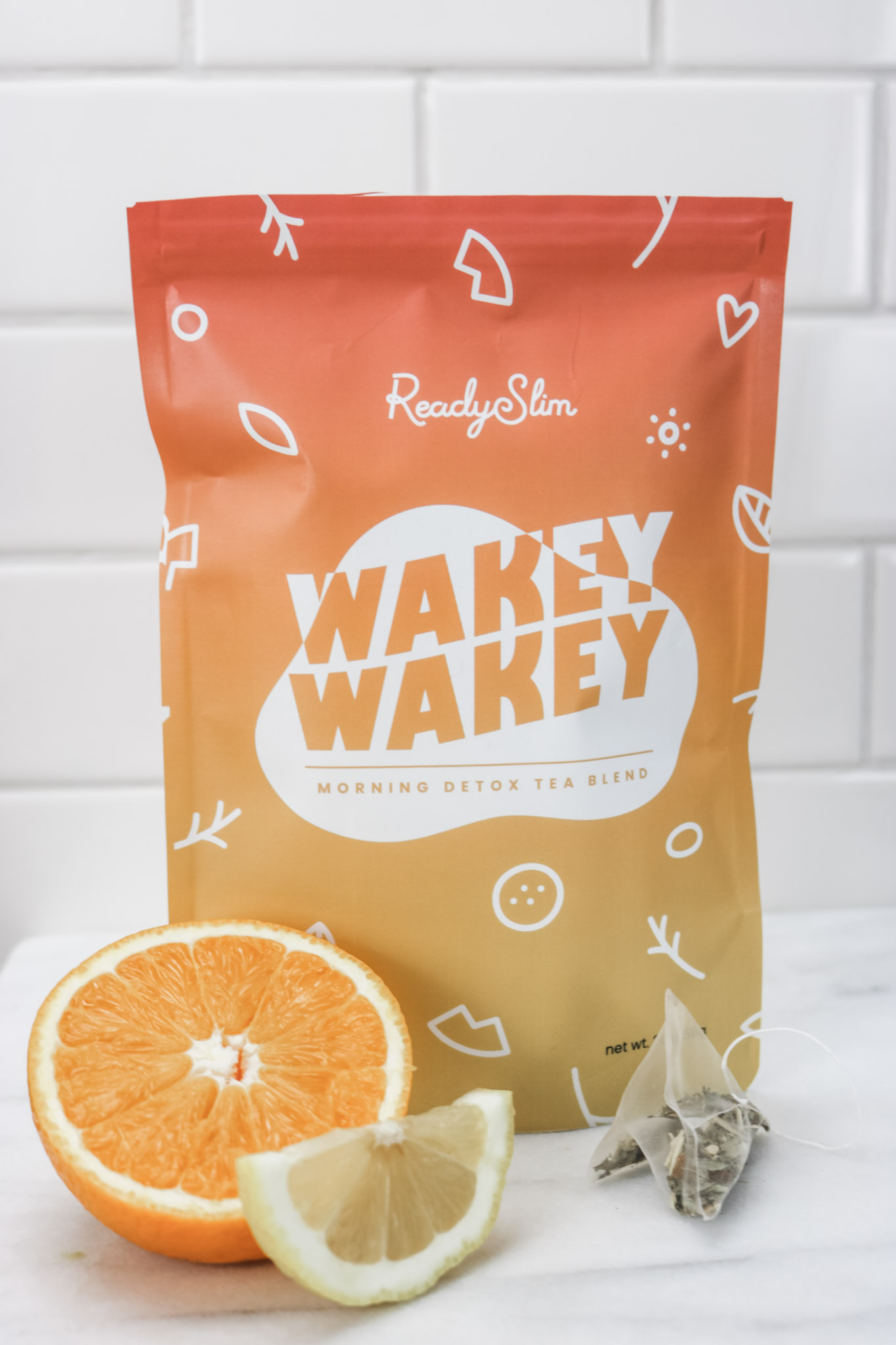 A bag of ReadySlim Wakey Wakey Morning Detox Tea Blend  is shown with a tea bag on the outside, and a slice of lemon and half an orange is shown. 