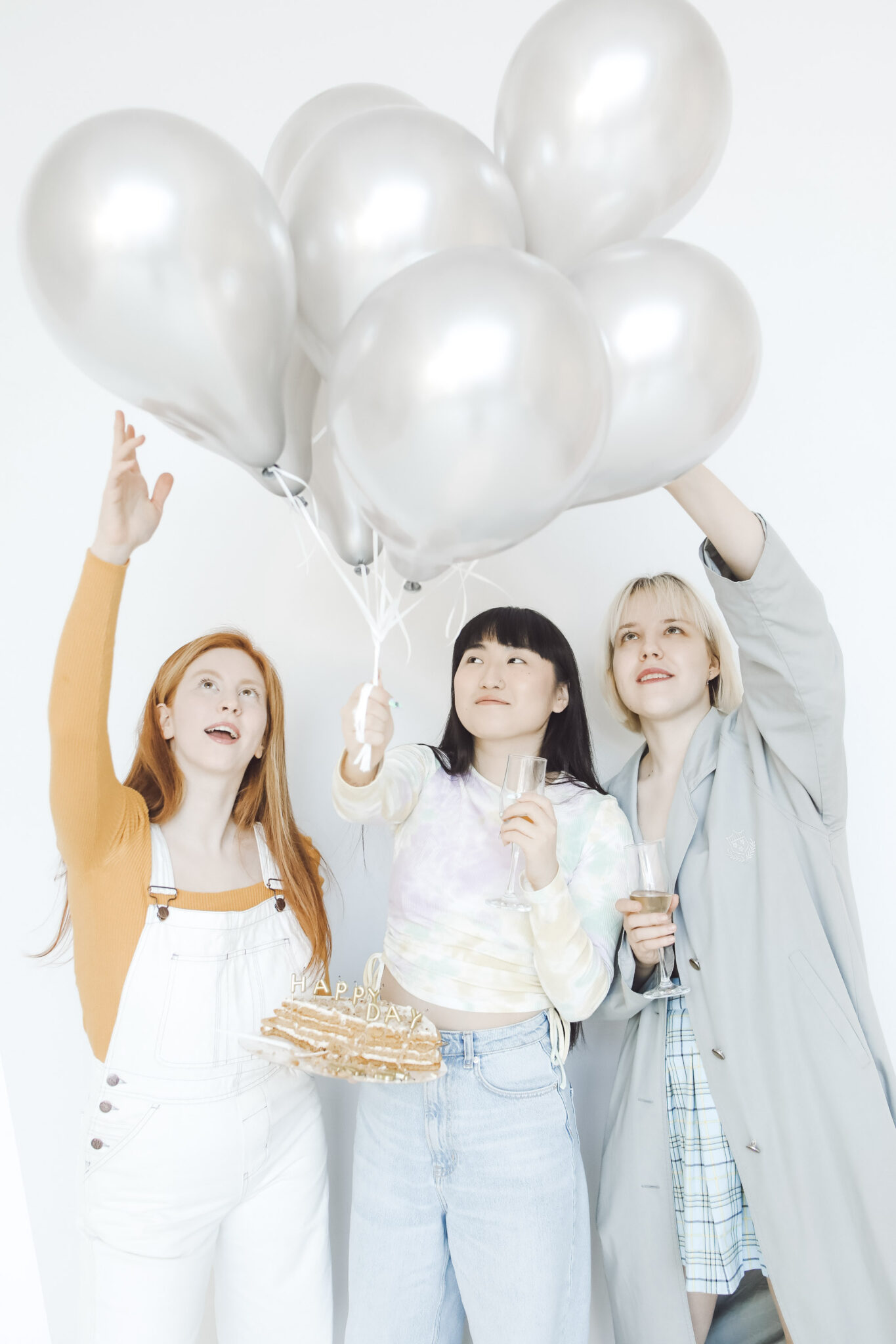 3 women hold silver balloons and look up. The middle woman holds her Birthday cake in surprise. This article covers how to give your best friend the best surprise Birthday party ever.