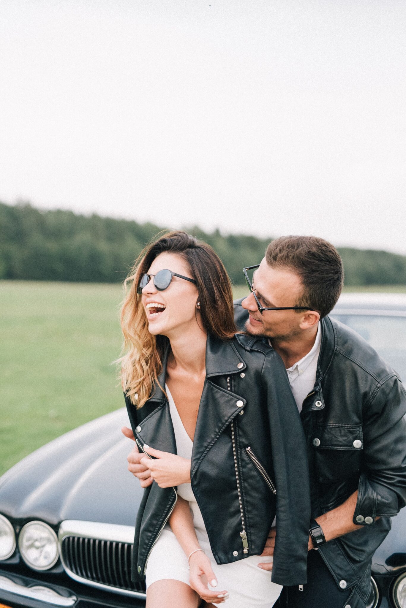 A man and woman sit on the hood of the car and laugh. They are on a trip. This article covers solutions for rented car problems.